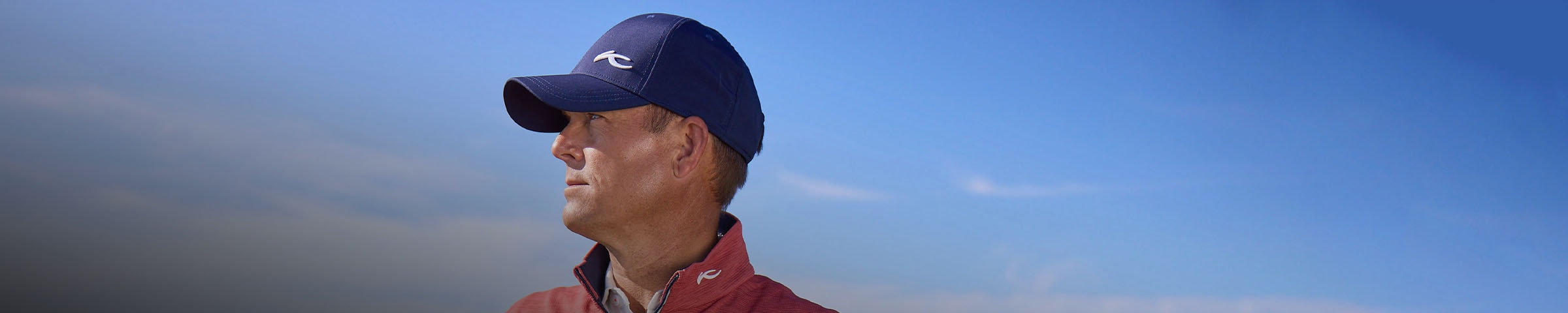 A golfer in profile wearing a KJUS golf cap looks over the course with the blue sky behind him.